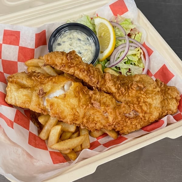 Golden-brown fried fish and french fries in a paper-lined container with tartar sauce, lemon, and salad.