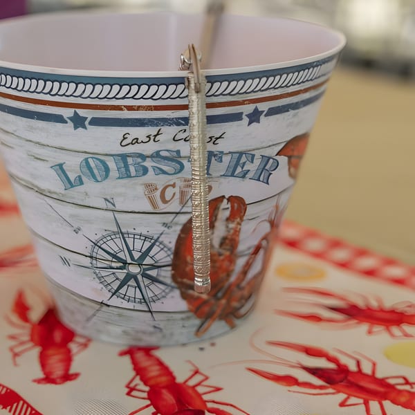 A white bucket with "East Coast Lobster" and a compass design, on a red lobster-patterned tablecloth.