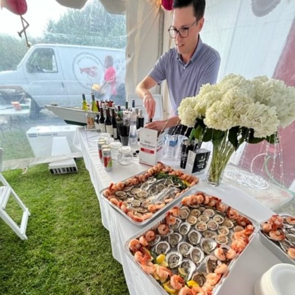 A man shucking oysters at an outdoor catering event with trays of seafood and drinks.