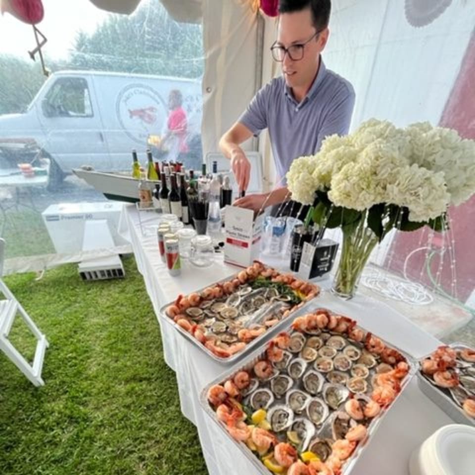 A man shucking oysters at an outdoor catering event with trays of seafood and drinks.