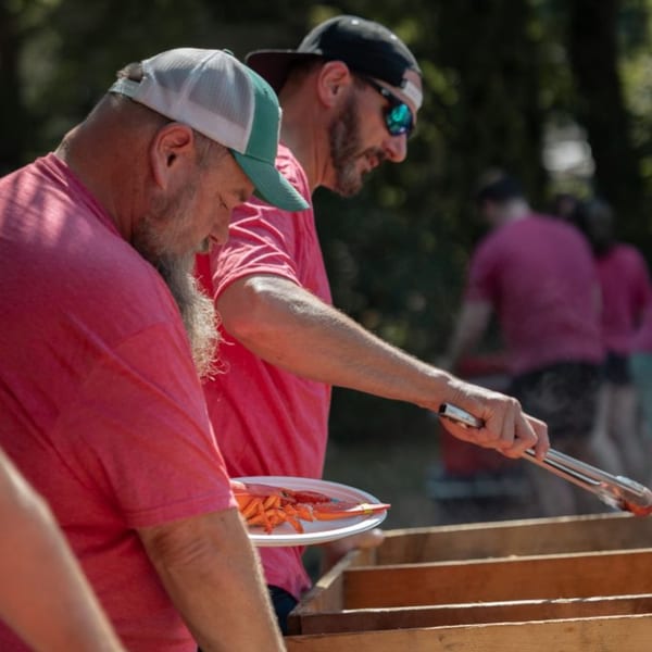 Two men in pink shirts and hats cooking lobster on a grill outdoors.
