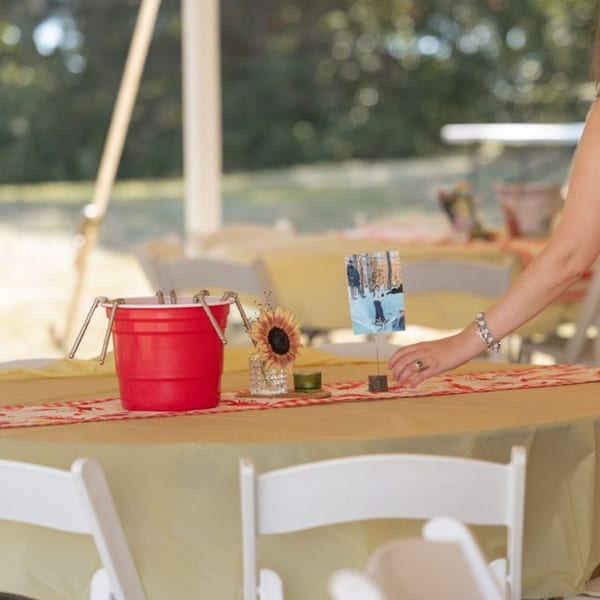 A woman's arm adjusts a table setting with a red bucket, sunflower, and photo.