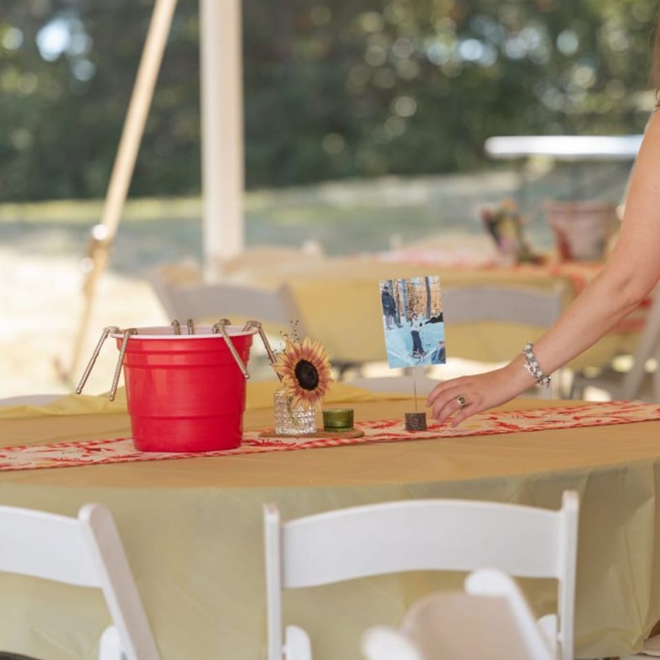 A woman's arm adjusts a table setting with a red bucket, sunflower, and photo.