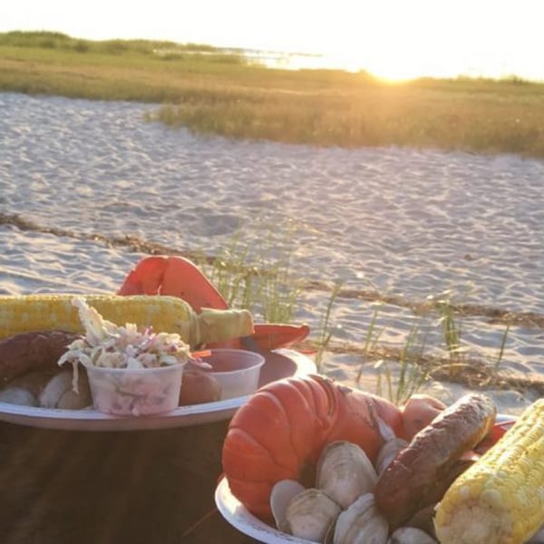 Lobster, clams, corn, and coleslaw on plates at a beachside sunset picnic.