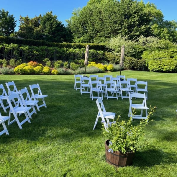 White chairs arranged on a lush green lawn for an outdoor event, surrounded by trees and shrubs.