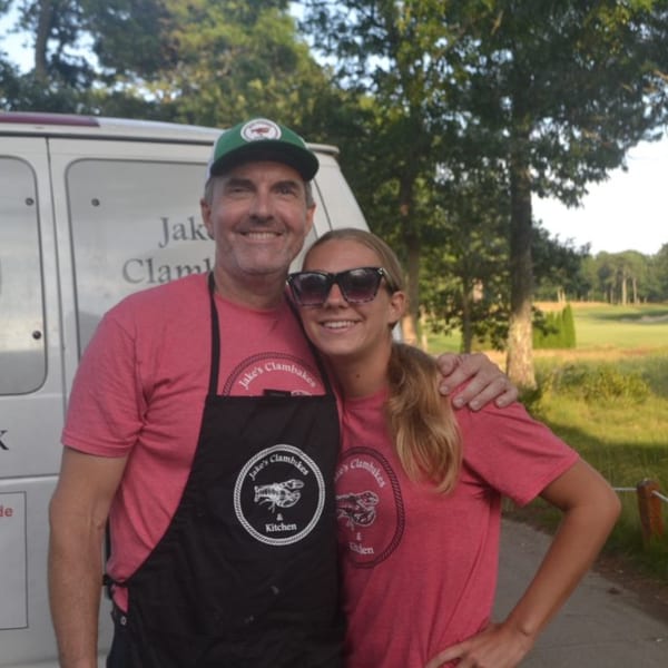 A man and woman in Jake's Clambakes shirts and apron smiling outdoors.