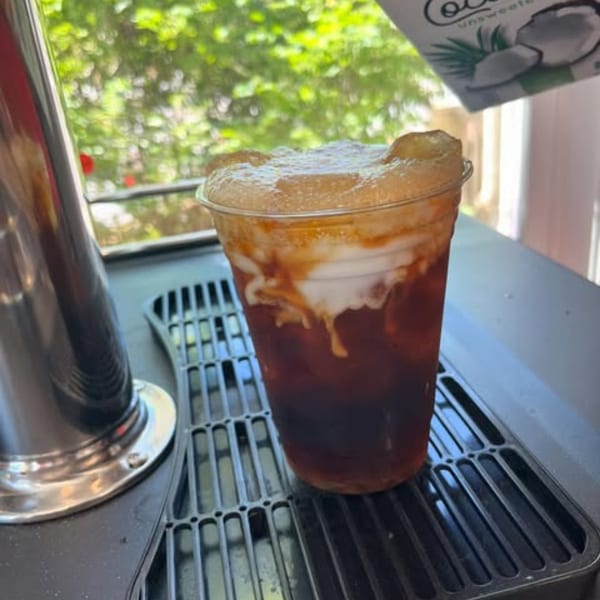 A plastic cup of iced coffee with white cream and foam on top, on a black grate.