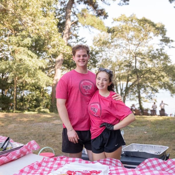 Two smiling people in pink shirts with a logo, standing behind a picnic table outdoors.