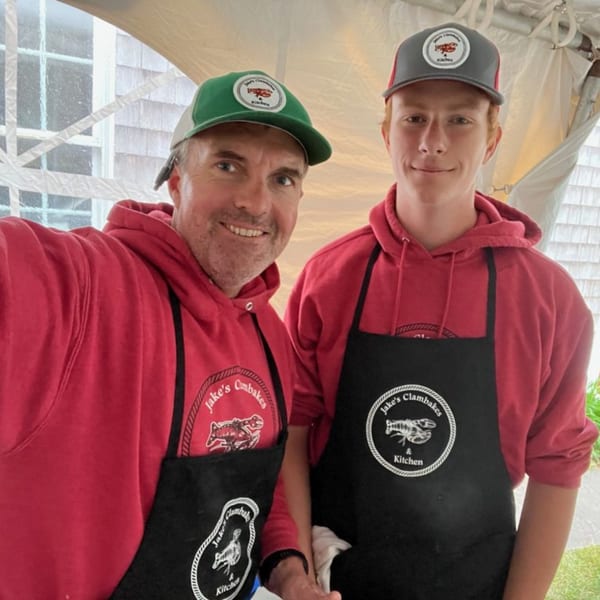 Two men in red hoodies and black aprons with "Jake's Clambakes & Kitchen" logos smile at the camera.
