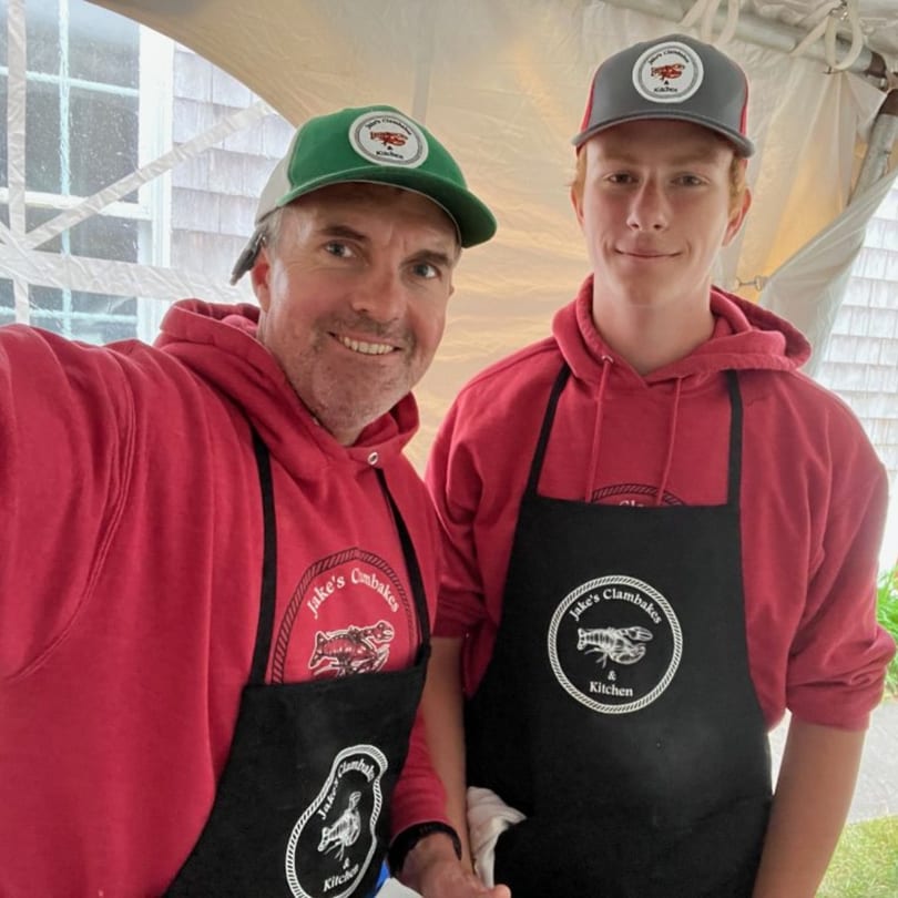 Two men in red hoodies and black aprons with "Jake's Clambakes & Kitchen" logos smile at the camera.