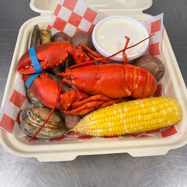 A lobster boil in a to-go container with lobster, clams, potatoes, corn, and dipping sauce.