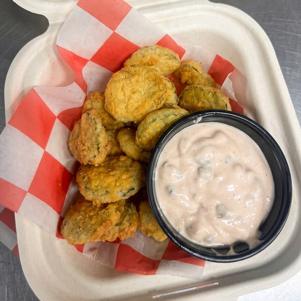 Golden-brown fried pickles in a paper container with red and white checkered lining, alongside a dipping sauce.