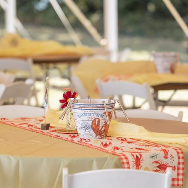 A table set with a yellow tablecloth and a lobster-themed runner, featuring a decorative bucket.