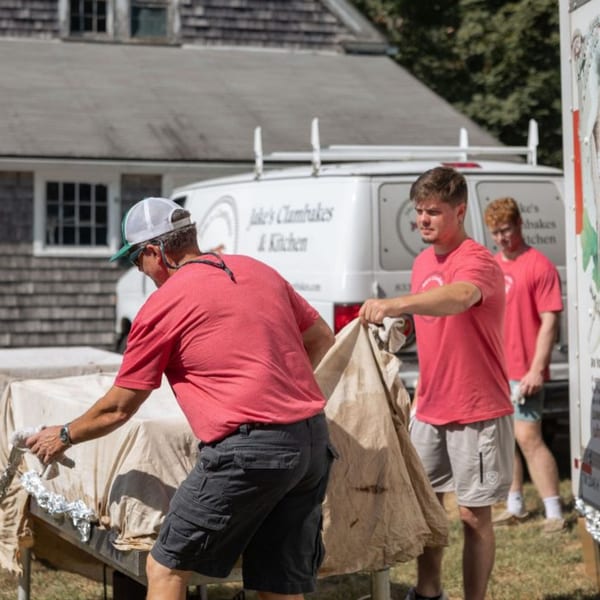Three men in red shirts and shorts setting up a table with a white van in the background.