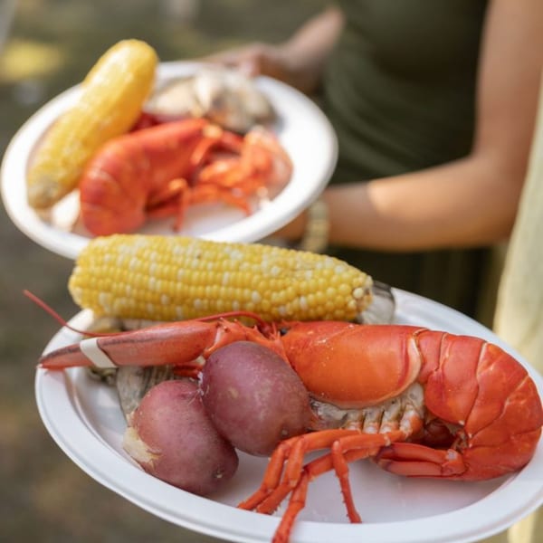 Two plates of lobster, corn on the cob, and potatoes are held by people.