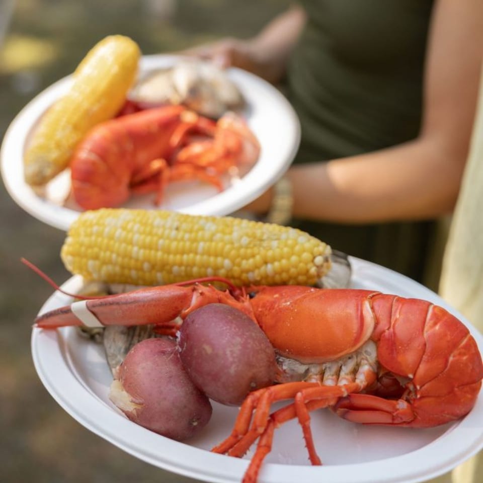 Two plates of lobster, corn on the cob, and potatoes are held by people.