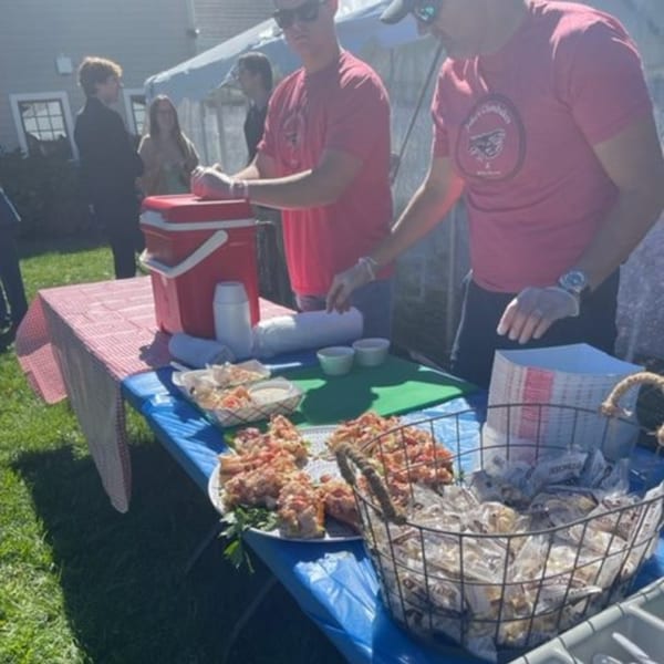 Two men in red shirts serving lobster rolls and other food from a blue and red checkered table outdoors.