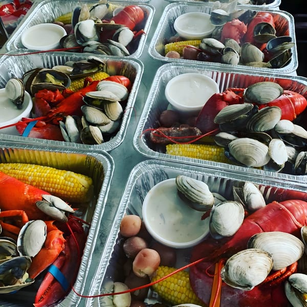 Multiple aluminum trays filled with lobsters, clams, corn on the cob, and potatoes.