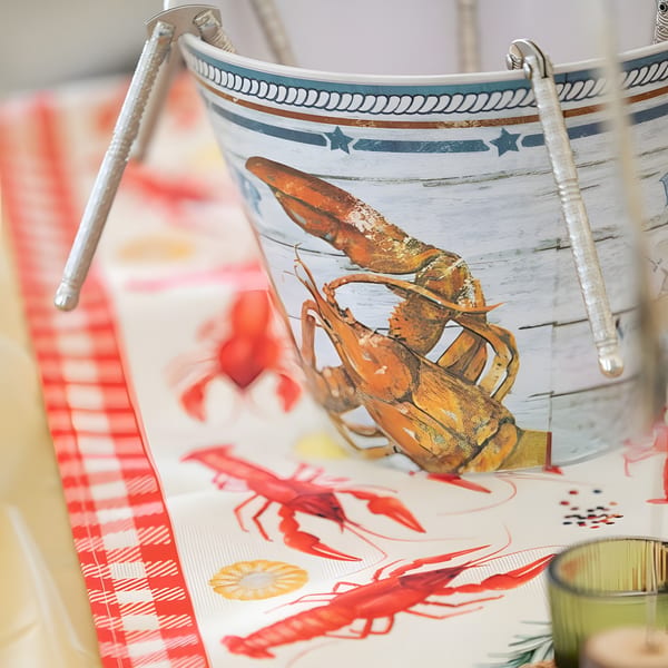 A bucket with a lobster design sits on a tablecloth with red lobster and checkered patterns.