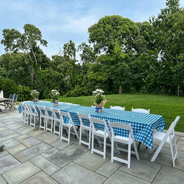 Long outdoor dining table with blue and white checkered tablecloths and white folding chairs on a patio.