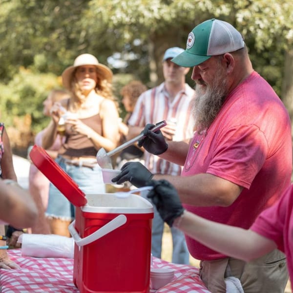 A man in a green hat and pink shirt serves food from a red cooler at an outdoor event.
