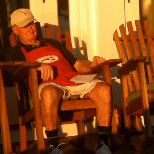 Man in a red apron and cap relaxing in a wooden Adirondack chair on a sunny porch.