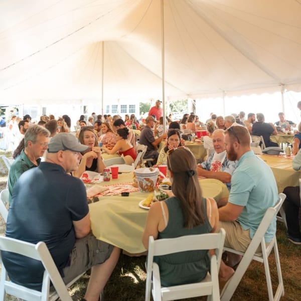 Guests seated at tables under a large white tent, enjoying a lively outdoor gathering.