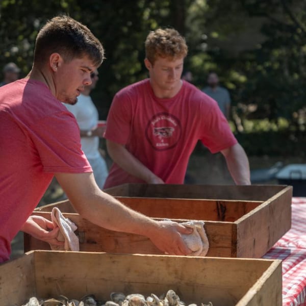 Two men in red shirts preparing oysters in wooden boxes on a red and white checkered tablecloth.