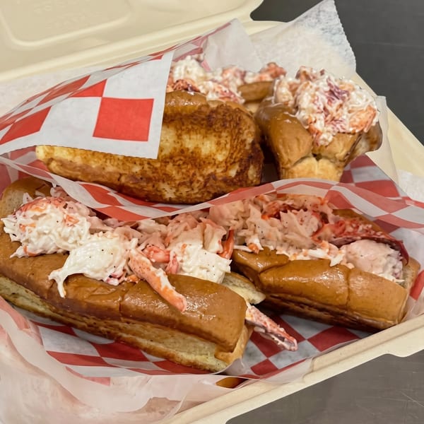 Four lobster rolls with creamy filling and red checkered paper in a takeout container.