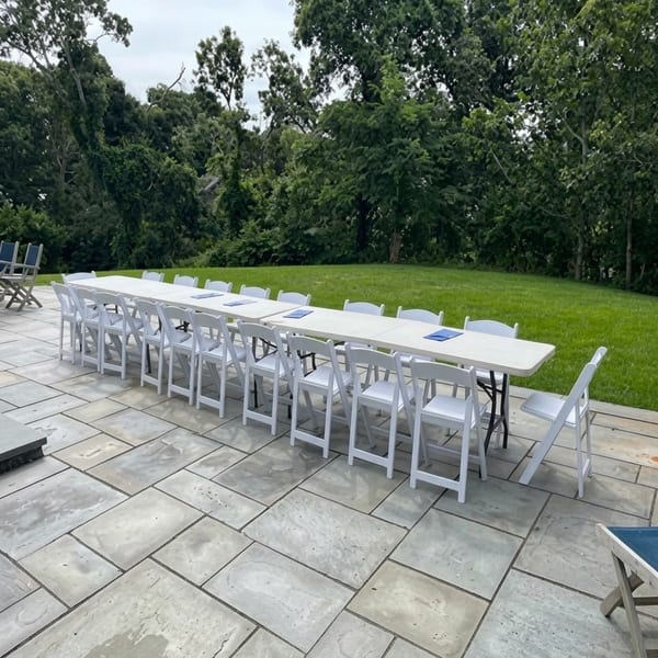 Long white tables and chairs set up on a patio for an outdoor event, surrounded by green grass and trees.