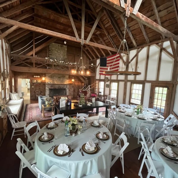 Rustic barn interior with wooden beams, set tables for an event, and a lounge area with a fireplace.