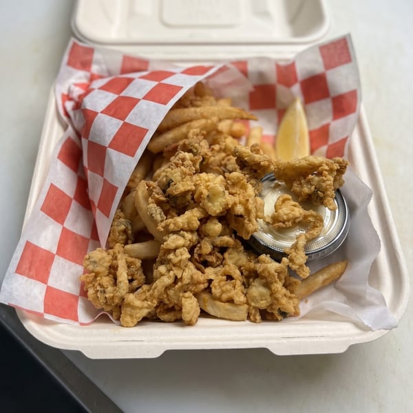 Fried clams and french fries in a takeout container with tartar sauce and a lemon wedge.