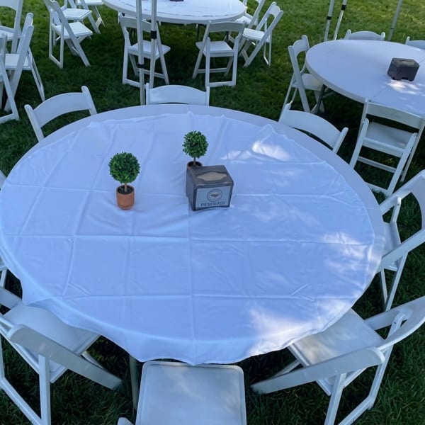 Outdoor event setup with white round tables, white chairs, and small potted plants on green grass.