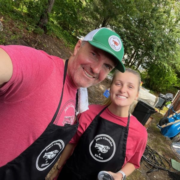 Two smiling people, a man and a woman, wearing black aprons with "Jake's Clambakes & Kitchen" logos.