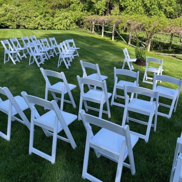 White folding chairs arranged on a vibrant green lawn for an outdoor event, with lush trees in the background.