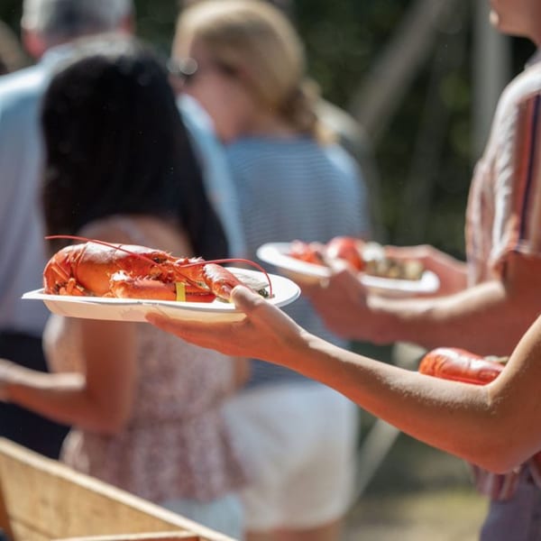 People holding plates with whole cooked lobsters at an outdoor gathering.