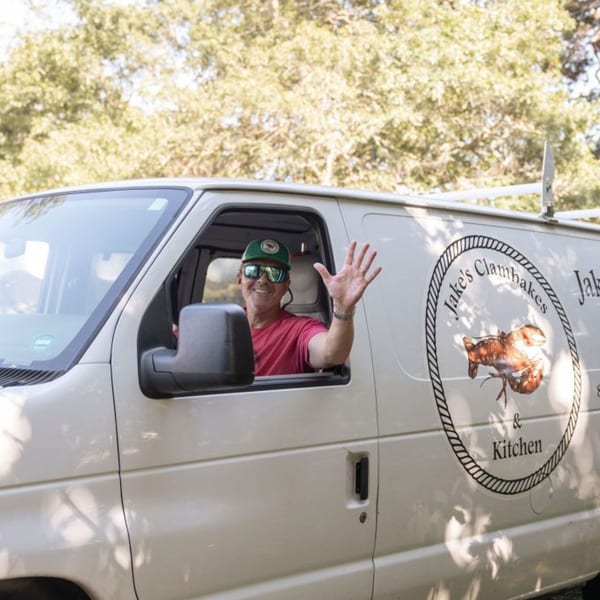 Man in sunglasses and hat waves from the driver's seat of a white Jake's Clambakes van.