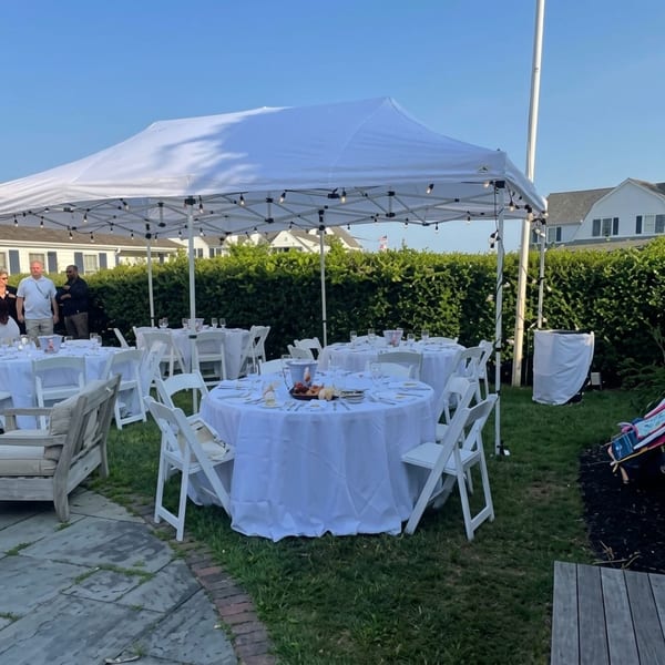 Outdoor event setup with white tents, round tables, chairs, and string lights on a sunny day.
