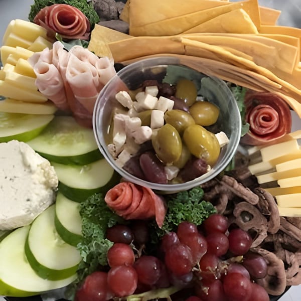 A charcuterie board with cheeses, meats, crackers, olives, grapes, cucumbers, and a white dip.