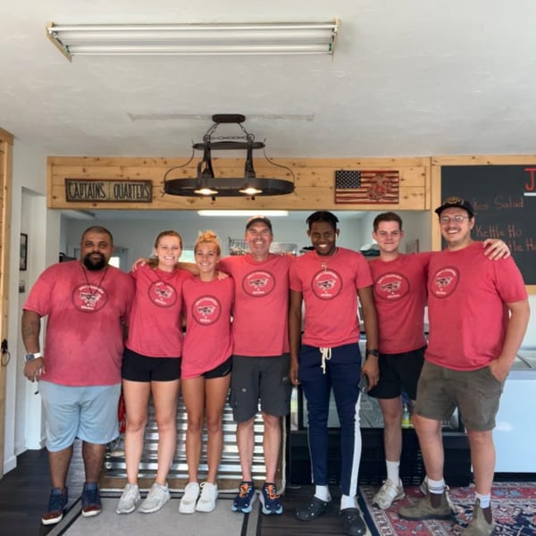 Six smiling restaurant staff members in red shirts stand together in a casual dining area.