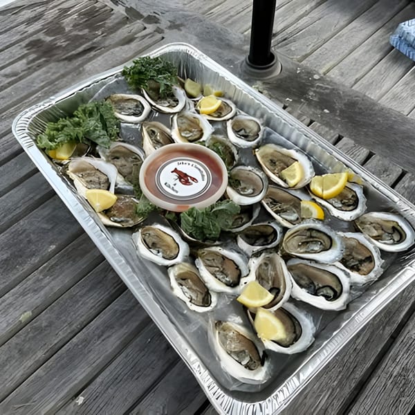 A foil tray of fresh oysters on ice with lemon wedges, parsley, and cocktail sauce on a wooden table.