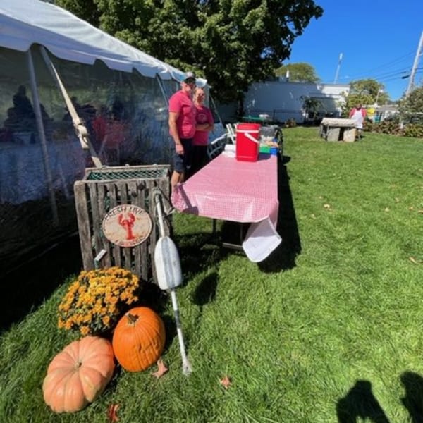 Two people stand by a red-checkered table with pumpkins and a lobster sign in a grassy yard.