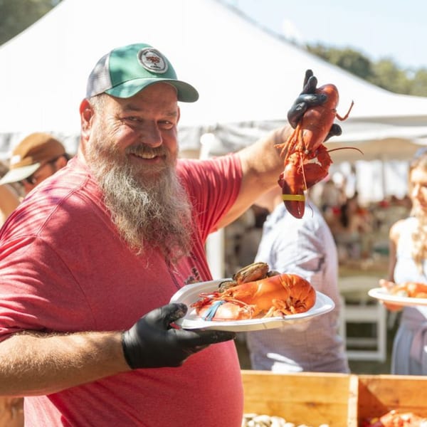 Smiling man in a green hat and black gloves holding up a cooked lobster and a plate of seafood.