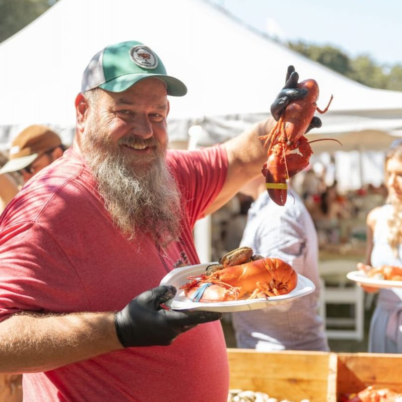 Smiling man in a green hat and black gloves holding up a cooked lobster and a plate of seafood.