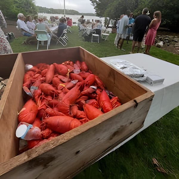 A wooden crate filled with cooked red lobsters, with people dining outdoors by a lake in the background.