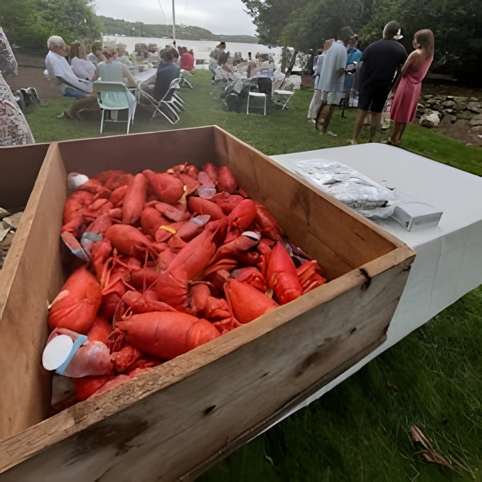 A wooden crate filled with cooked red lobsters, with people dining outdoors by a lake in the background.