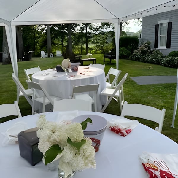 Outdoor event setup with white tables and chairs under a tent, surrounded by green lawn and trees.
